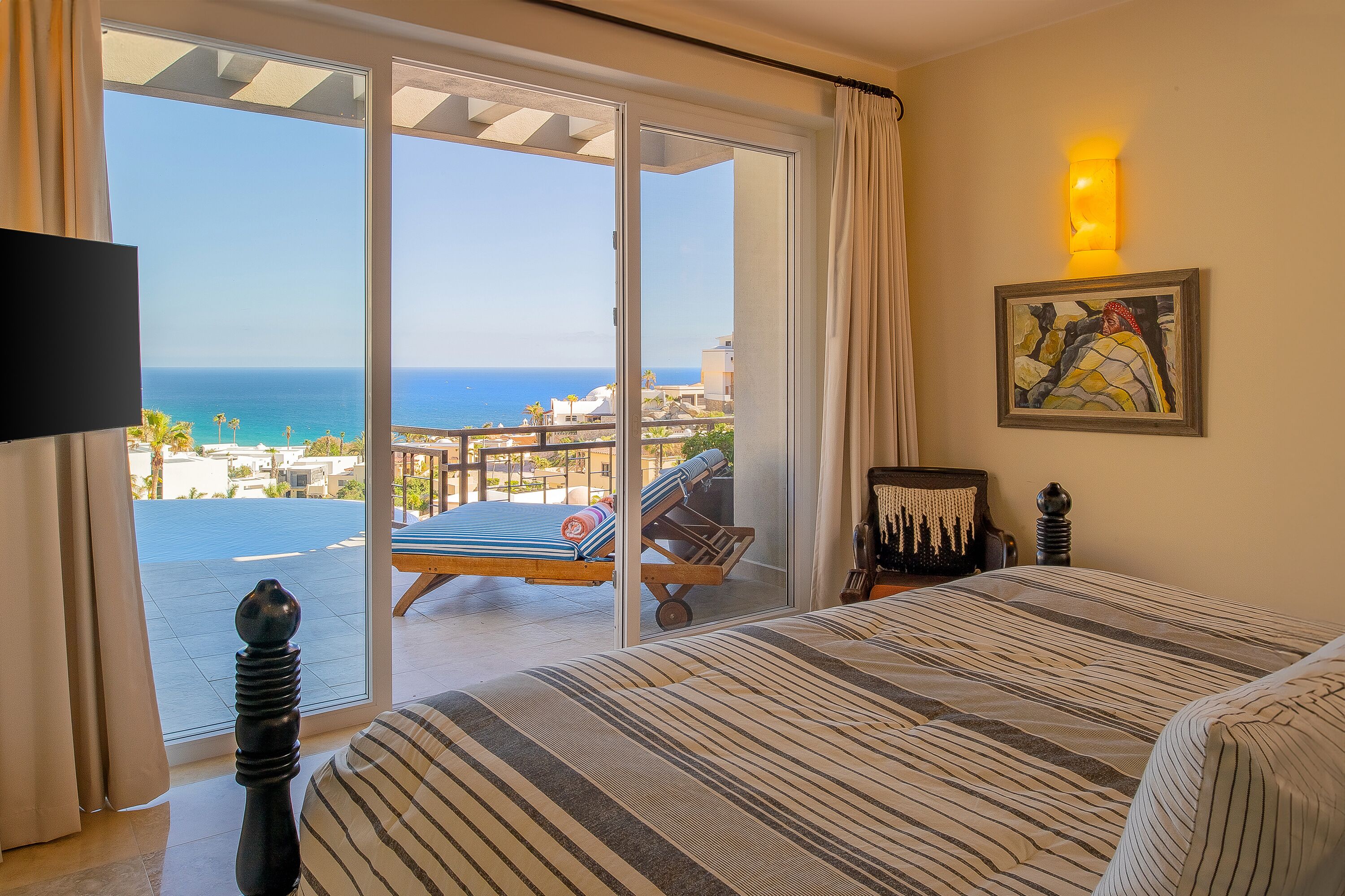 Hotelkamer met een grote schuifglazen deur naar een balkon met uitzicht op zee. Er is een bed met gestreepte lakens, een stoel met een kussen en een kleine tafel met een lamp.