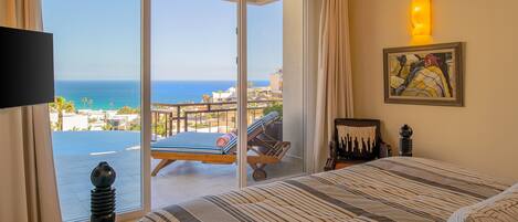 Hotelkamer met een grote schuifglazen deur naar een balkon met uitzicht op zee. Er is een bed met gestreepte lakens, een stoel met een kussen en een kleine tafel met een lamp.
