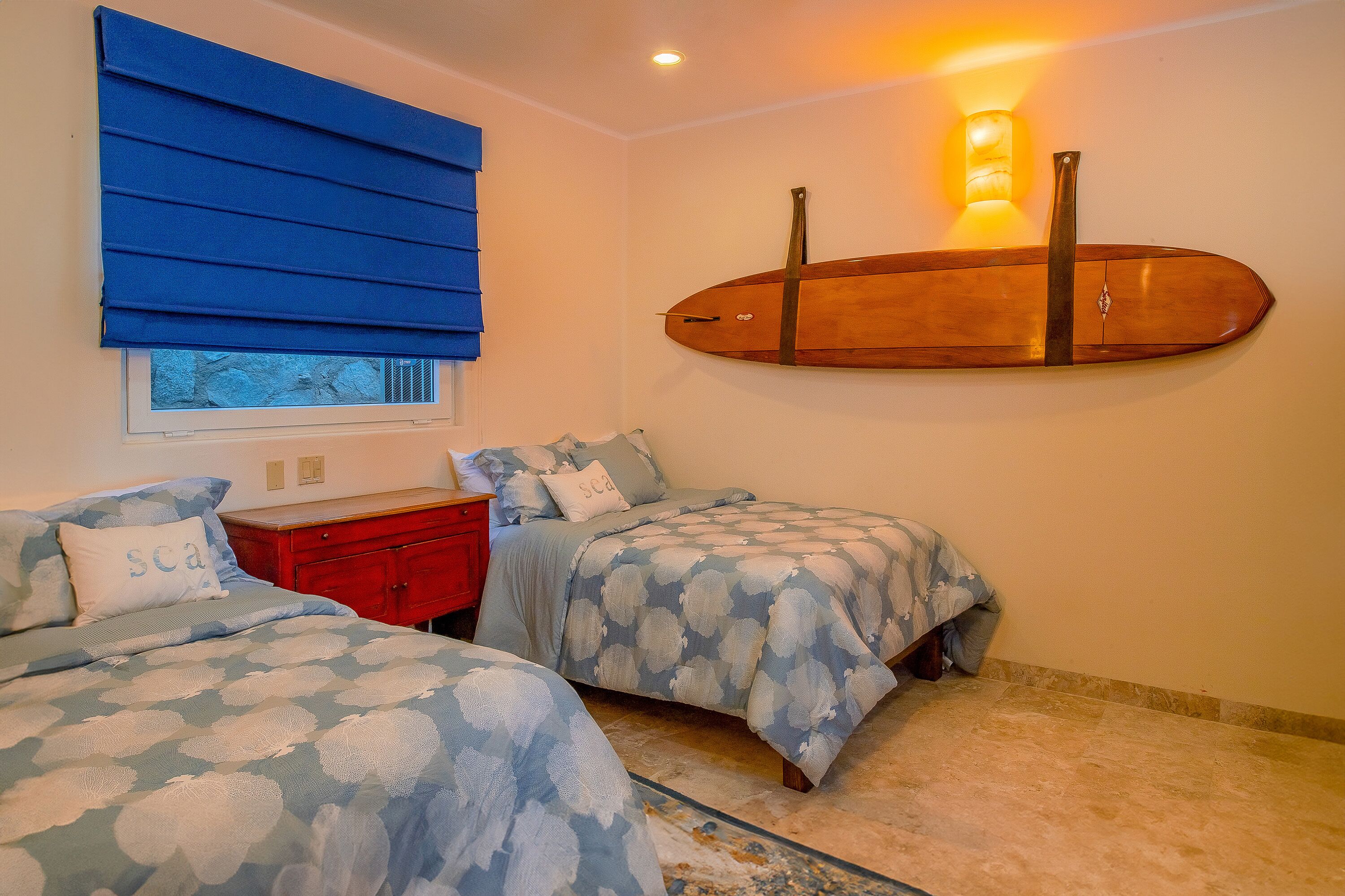 Una camera da letto con due letti, una finestra con persiane blu, un'asse da surf appesa al muro e un comodino con una lampada.