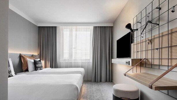 Habitació d'hotel moderna amb dos llits, un finestral gran amb cortines, un escriptori de fusta i un televisor penjat.