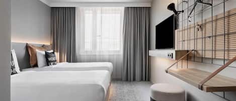 Une chambre d’hôtel moderne avec deux lits, une grande fenêtre avec des rideaux, un bureau en bois et un téléviseur fixé au mur.
