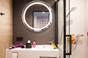 A modern bathroom with a round mirror, a white sink, a hairdryer, and a shower with a colorful flower decoration. - Moxy Vienna City East (Vienna)