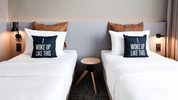A hotel room with two beds, each with a pillow that reads "Woke up like this." There is a small round table between the beds. - Moxy Vienna City East (Vienna)
