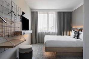 A modern hotel room with a bed, a TV mounted on a wall, a small table, and a bench. - Moxy Vienna City East (Vienna)