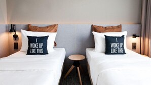 Una habitació d'hotel amb dos llits, cadascun amb un coixí que diu "Woke up like this". Hi ha una taula rodona petita entre els llits.
