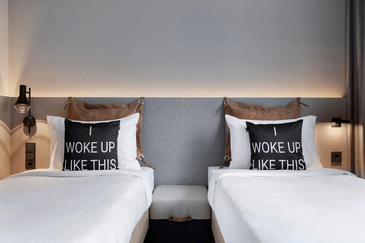 Une chambre d’hôtel avec deux lits, chacun agrémenté d’un oreiller sur lequel est brodé le message : « Je me suis réveillé(e) comme ça. »