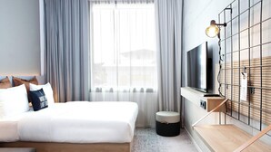 A modern hotel room with a large bed, a TV mounted on a wooden panel, a small stool, and a window with curtains. - Moxy Vienna City East (Vienna)
