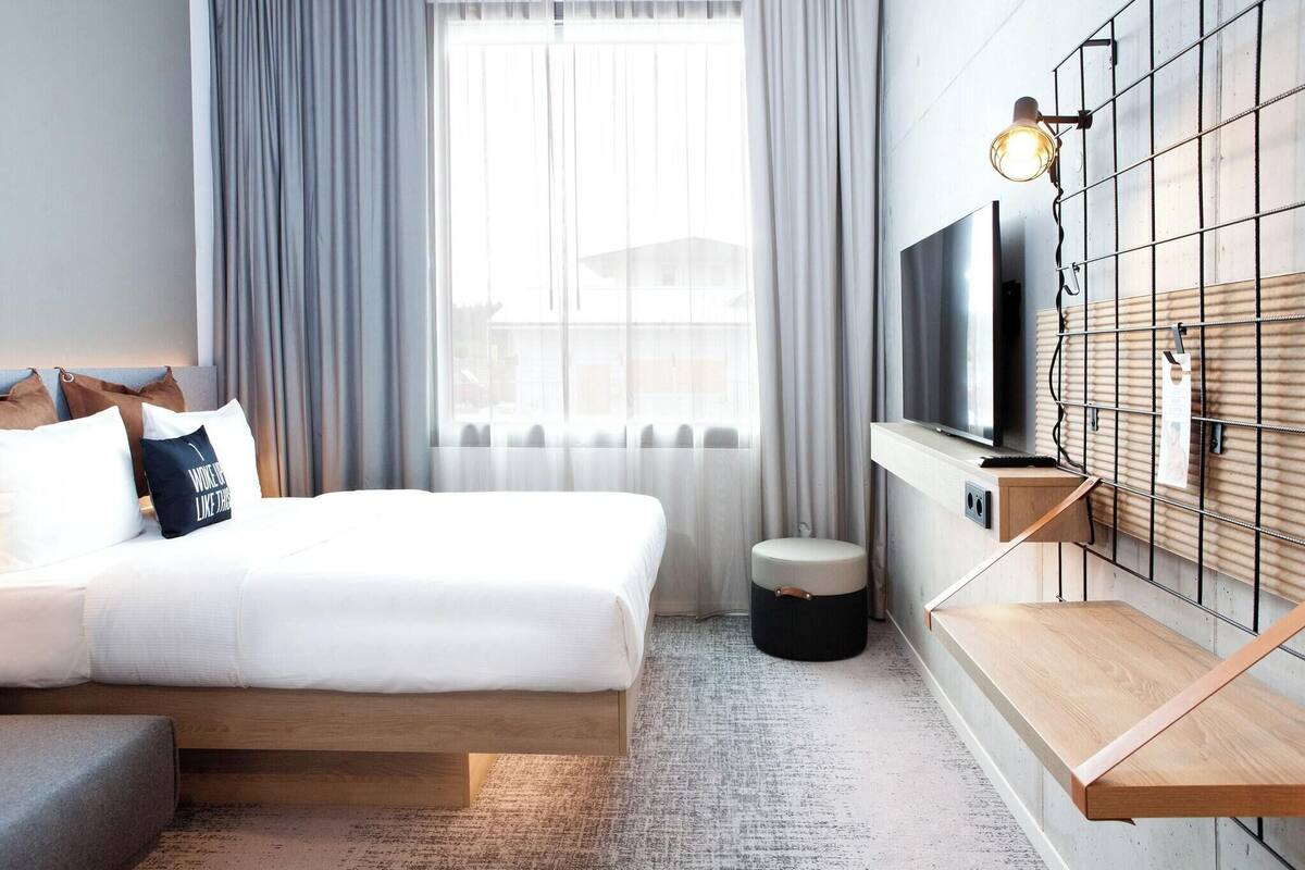 A modern hotel room with a large bed, a TV mounted on a wooden panel, a small stool, and a window with curtains.