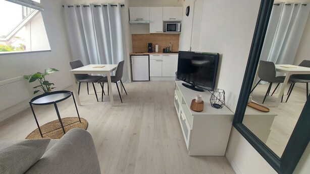 A modern, well-lit living room with a sofa, dining table, and chairs. A kitchen area with a microwave and cabinets is visible in the background. - Studio Cosy Le 12 (Châlons-en-Champagne)