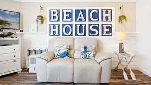 Una sala de estar con un sofá color beige, dos cojines decorativos, un letrero en la pared que dice "BEACH HOUSE" y una pequeña mesita de noche con una lámpara.