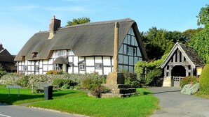 A traditional half-timbered house with a thatched roof, a stone cross, and a small wooden structure. - Inviting 3-bedroom Lodge in Ashton Under Hill (Evesham)