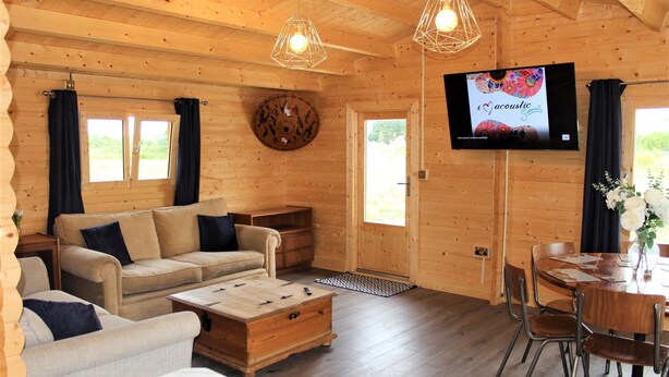 A wooden cabin interior with a sofa, coffee table, dining table, chairs, a TV, and a window with curtains. - Inviting 3-bedroom Lodge in Ashton Under Hill (Evesham)