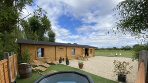 A wooden building with a patio area, a hot tub, and a grassy field in the background. - Inviting 3-bedroom Lodge in Ashton Under Hill (Evesham)