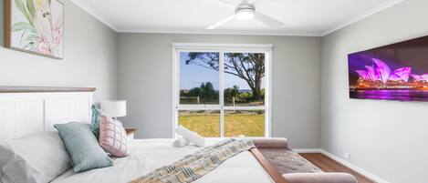 A bedroom with a bed, a bench, a ceiling fan, a window with a view of a tree and grass, and a painting of a bird on the wall.