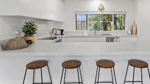 Une cuisine moderne avec des meubles blancs, un grand îlot central doté de tabourets, une fenêtre donnant sur des palmiers et une horloge au mur.