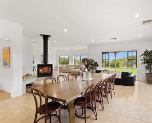 A spacious living area with a large dining table, comfortable seating, a fireplace, and large windows offering a view of the outdoors.