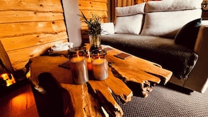 Ein gemütliches Zimmer mit einer Wand aus Holzstämmen, einem Holztisch mit Kerzen und einer Vase, einem grauen Sofa und einer kleinen Pflanze.