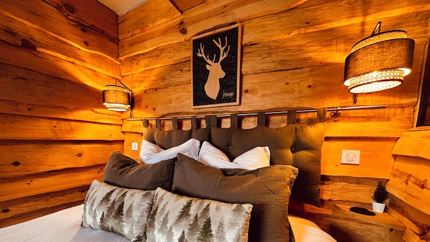 Ein gemütliches Schlafzimmer in einer Blockhütte mit einem Bett, Kissen, einem Kopfteil, Wandlampen und einer Hirsch-Silhouette als Bild.
