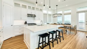 A modern kitchen with white cabinetry, a large island with bar stools, pendant lighting, and a view to the outside. - Private Beachfront Home w/ 5 Decks, 3 Suites, Fenced Yard in Beachside Village (Galveston)