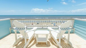 A balcony with two white Adirondack chairs and a small table, overlooking a beach with a clear sky and a bird flying. - Private Beachfront Home w/ 5 Decks, 3 Suites, Fenced Yard in Beachside Village (Galveston)