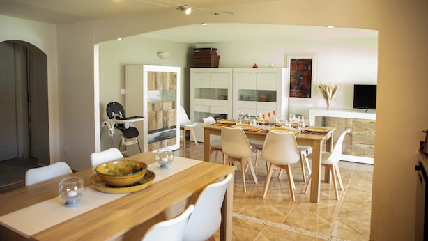 Un espace repas moderne avec une table en bois et des chaises blanches, une cuisine en arrière-plan et un téléviseur fixé au mur.