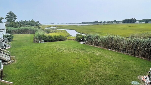 A grassy area with a small water body, a wooden deck, and a distant view of a house and fields. - Captain's Quarters -Water Front- Water Access-Pet Friendly-NO LINENS (Chincoteague)