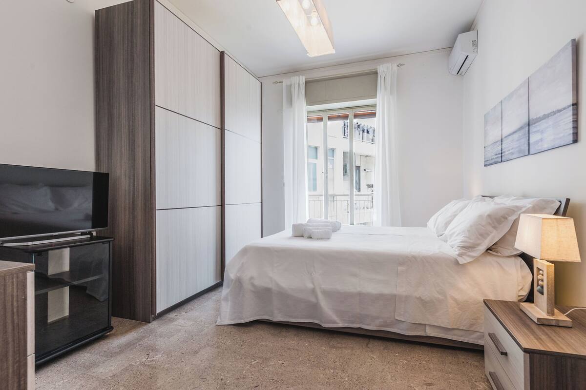 A modern hotel room with a bed, a TV, a bedside table with a lamp, and a view of the city through the window.