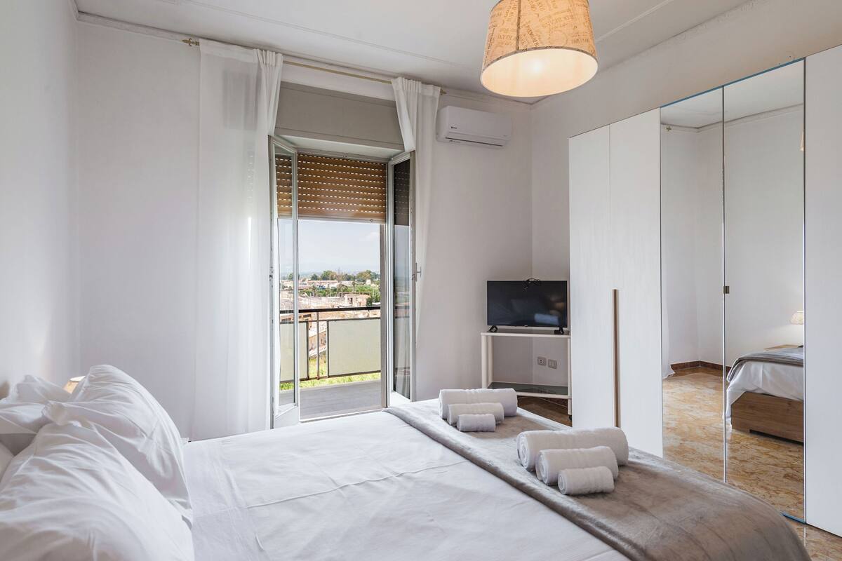 A modern hotel room with a large bed, a TV, and a balcony with a view of the city.