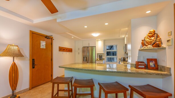 A modern kitchen with a central island, bar stools, a refrigerator, and a Buddha statue. - Royal Mauian #219 2 Bedroom Condo by RedAwning (Kihei)