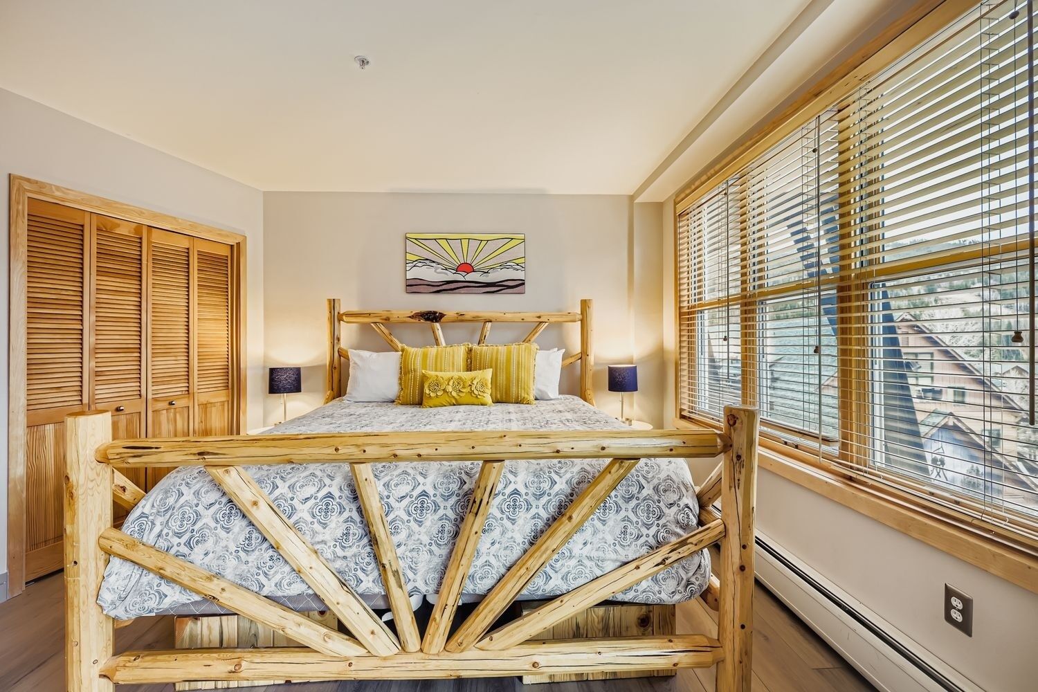 A room with a wooden bed frame, a large window with blinds, and a painting on the wall.