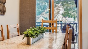 Un comedor con mesa de madera, sillas, planta en maceta y un amplio ventanal con vista a un paisaje boscoso.