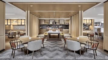 Uma zona de refeições moderna num hotel, com uma mesa central rodeada por cadeiras, uma cozinha ao fundo e um chão com padrão.