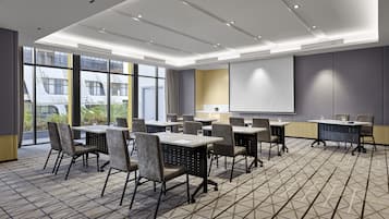 Una sala de conferencias moderna con mesas, sillas, una pantalla de proyector y ventanas amplias que ofrecen vistas al exterior.