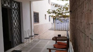 Un estrecho pasillo exterior con suelo de baldosas, sillas de metal, una mesa de madera con cojines anaranjados y una verja decorativa de metal.