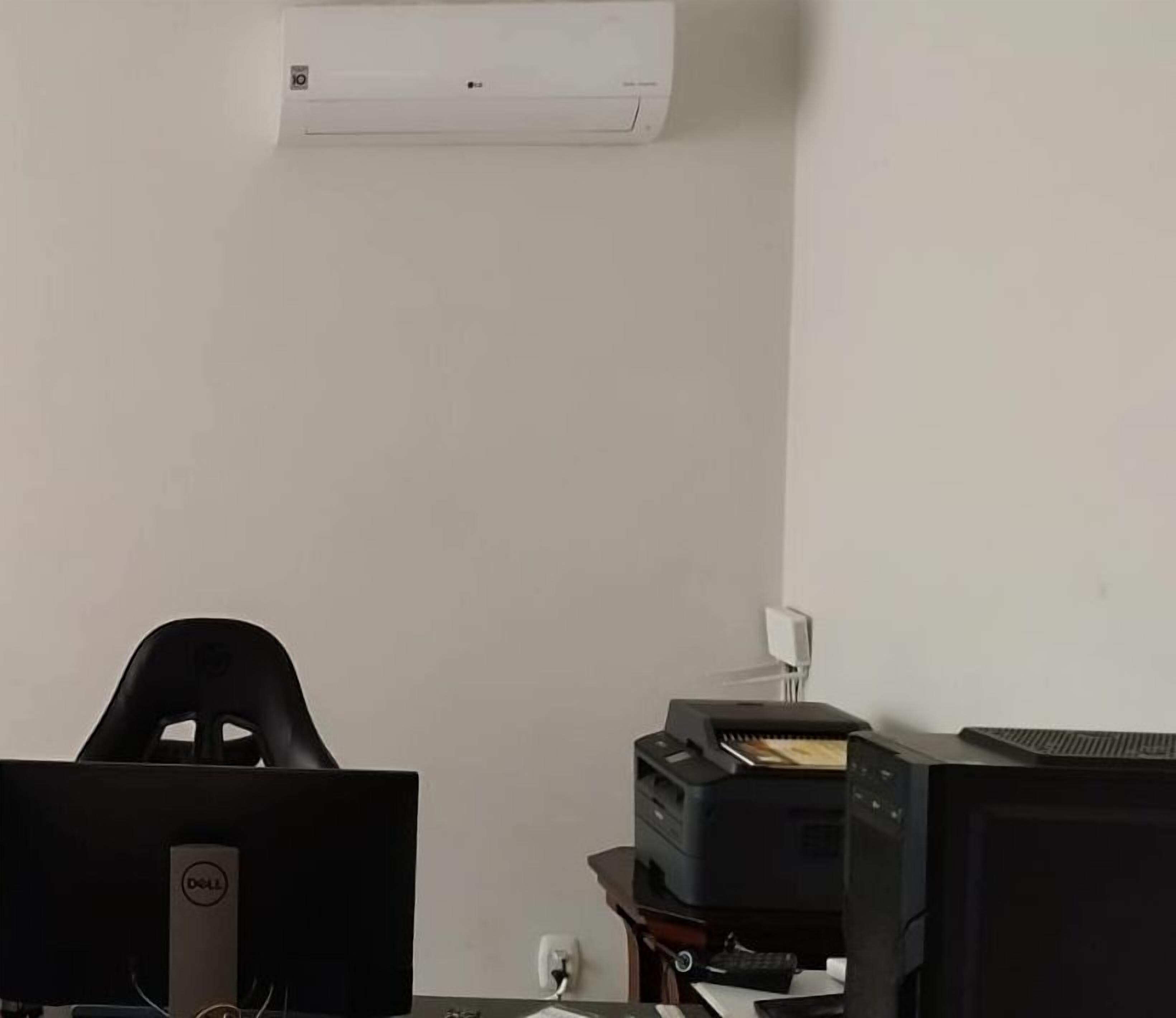 Quarto com escrivaninha, computador, impressora e ar condicionado fixado na parede.