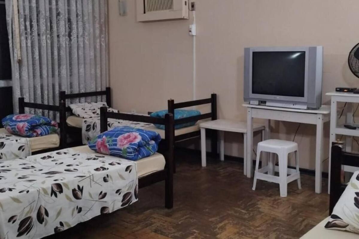 A room with two single beds, a television on a stand, a small white stool, and a window with curtains.