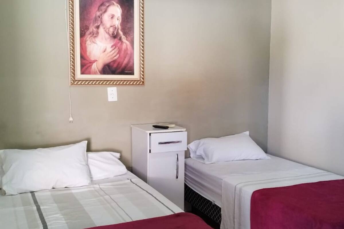 A room with two beds, each with white bedding and a red blanket on one bed. There is a small white cabinet with a remote control on top. A framed picture hangs on the wall above the cabinet.