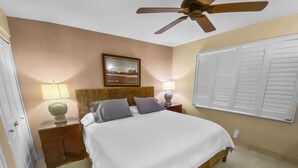 A hotel room with a bed, a ceiling fan, a nightstand with a lamp, a picture on the wall, and a window with shutters. - Maalaea Surf #g-2 1 Bedroom Condo by RedAwning (Kihei)