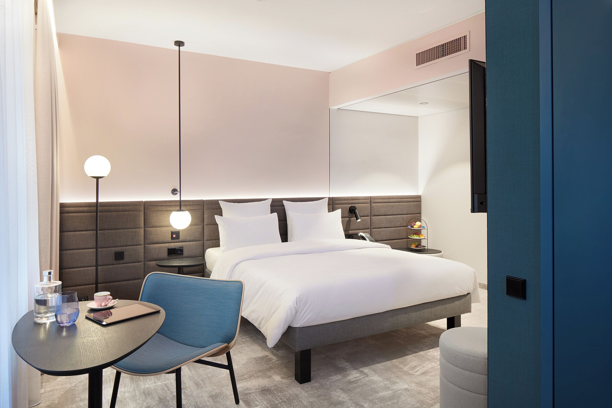 A modern hotel room with a bed, bedside table, chair, and a TV mounted on the wall.