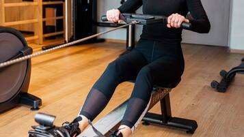 A person using a rowing machine in a gym with wooden flooring and exercise equipment in the background.