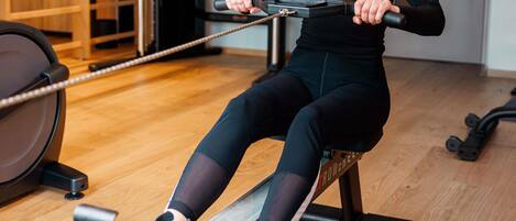 Una persona usando una máquina de remo en un gimnasio con suelo de madera y equipo de ejercicio al fondo.