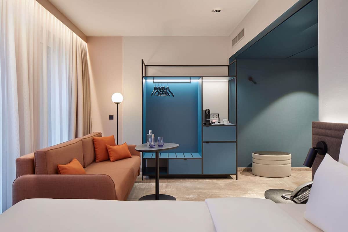 A modern hotel room with a bed, a sofa, a small table with glasses, a coffee machine, and a phone.