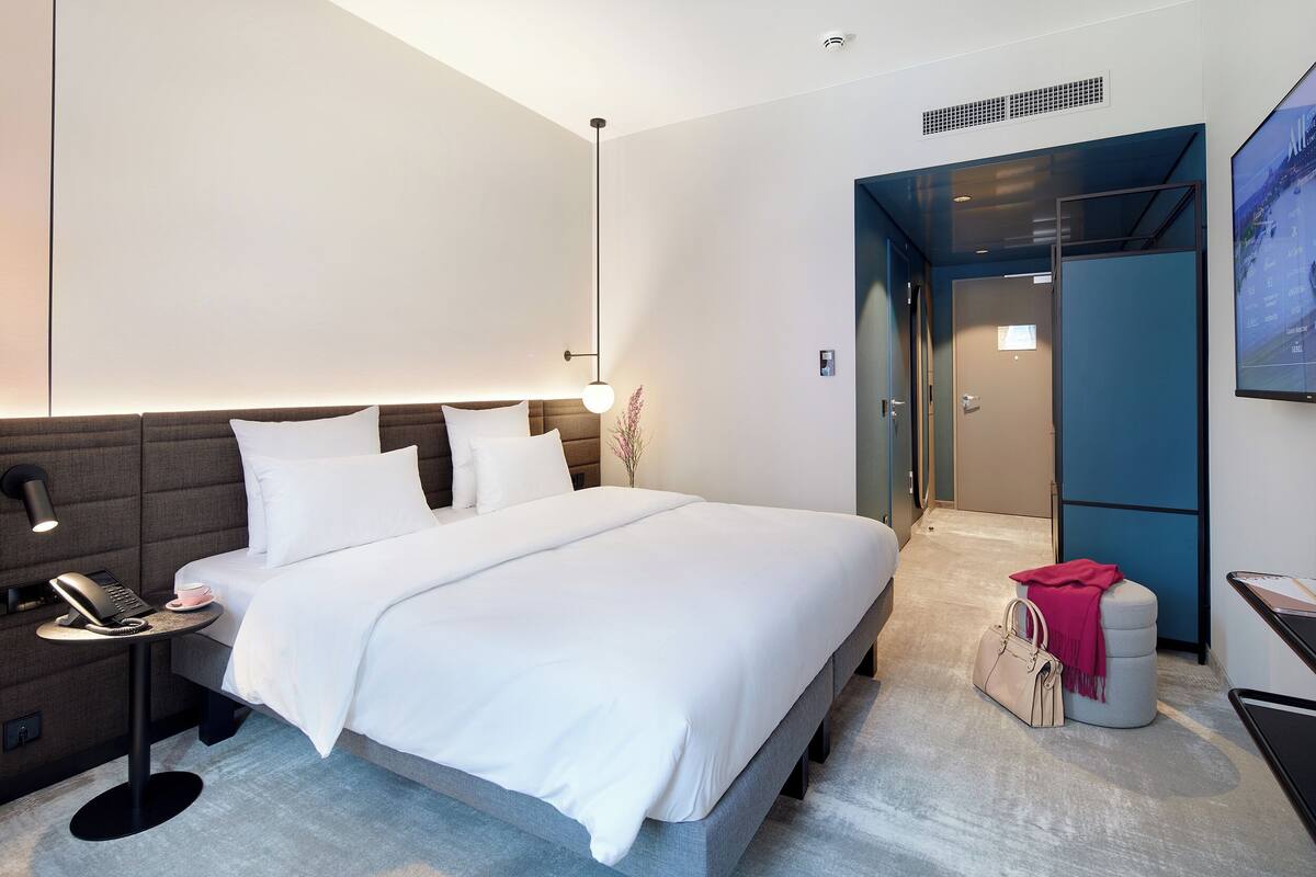Une chambre d’hôtel moderne avec un grand lit, une table de chevet avec une lampe, un téléphone et une petite table avec une tasse. On aperçoit une valise et une couverture rose sur un banc.