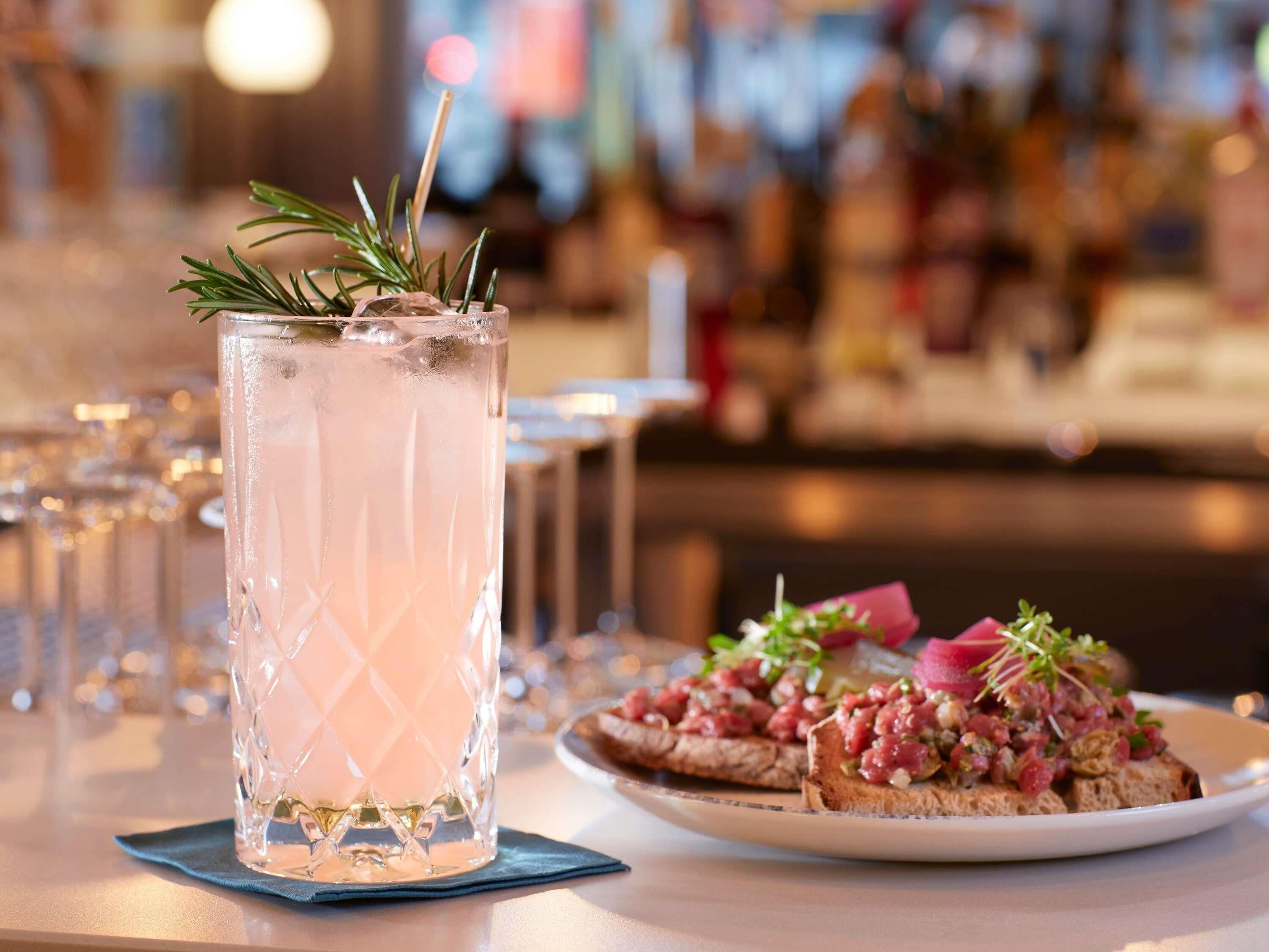 A tall glass with a clear liquid and a sprig of rosemary, a plate with a food item, and a bar setting in the background.