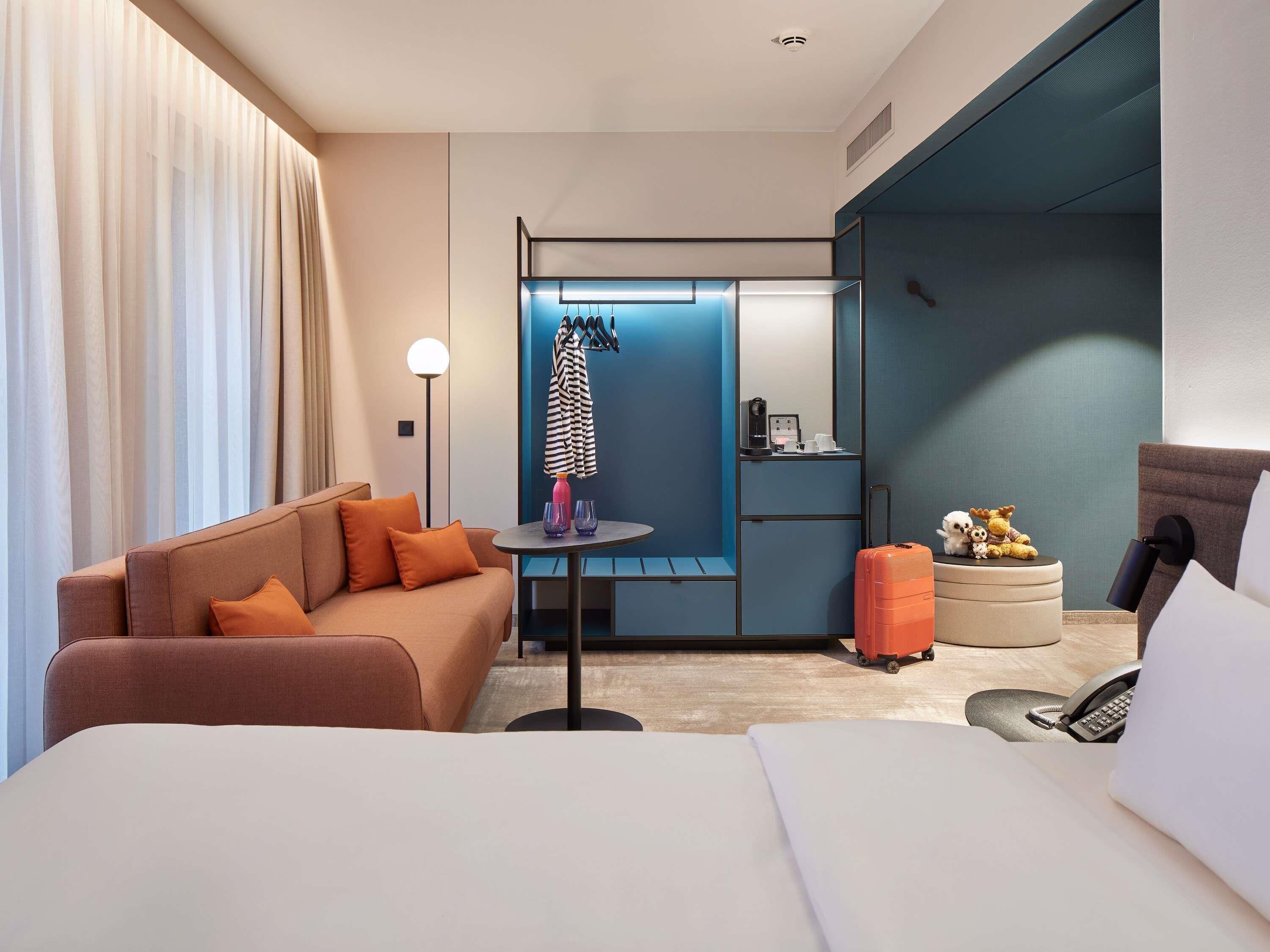 Ein modernes Hotelzimmer mit Bett, einer Couch, einem kleinen Tisch mit Lampe, einem Koffer und einer Minibar.