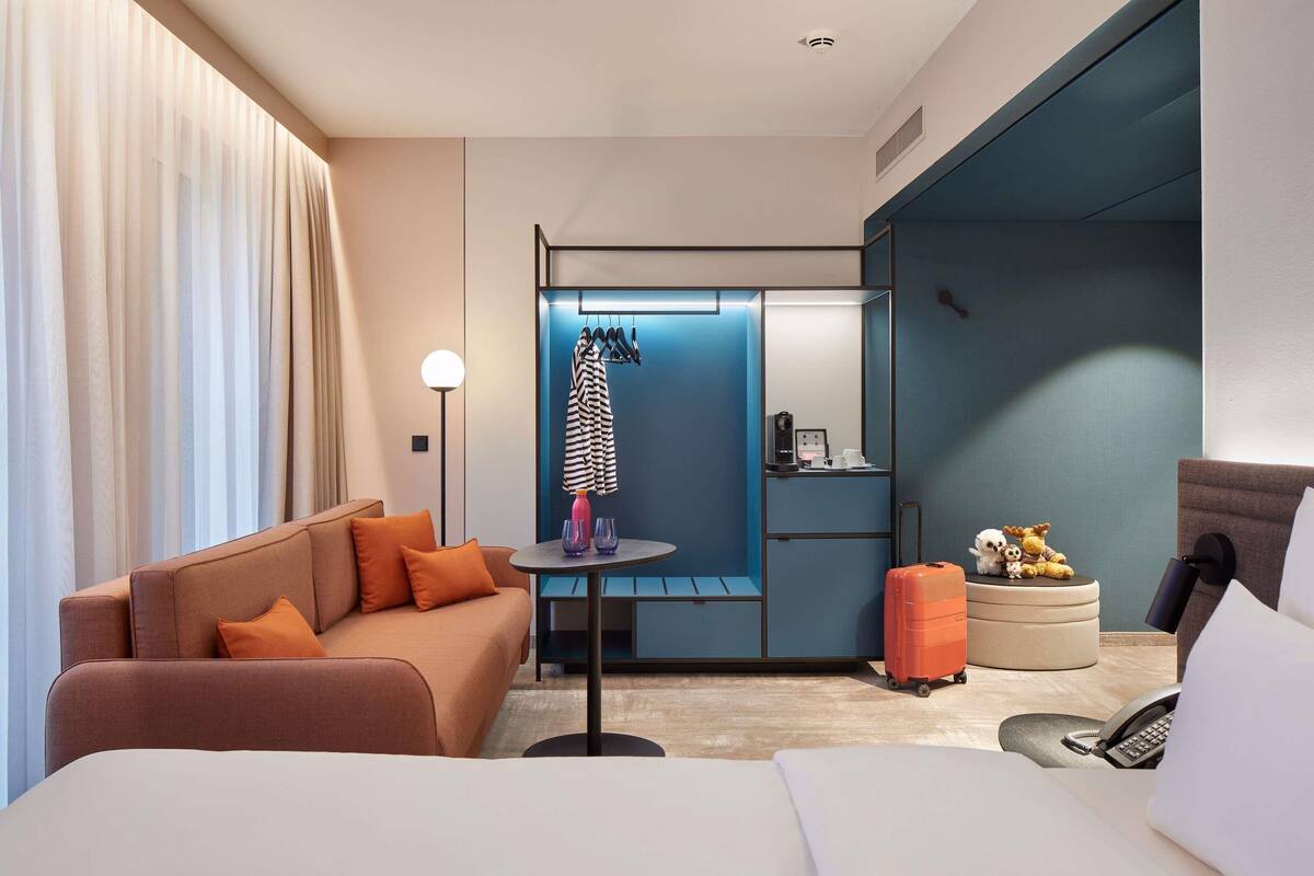 Une chambre d’hôtel moderne équipée d’un lit, d’un canapé, d’une petite table avec une lampe, d’une valise et d’un mini-bar.