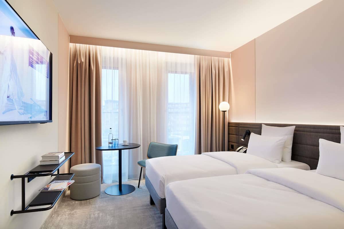 Une chambre d’hôtel avec deux lits, une petite table avec une chaise, un téléviseur fixé au mur et une fenêtre avec des rideaux.