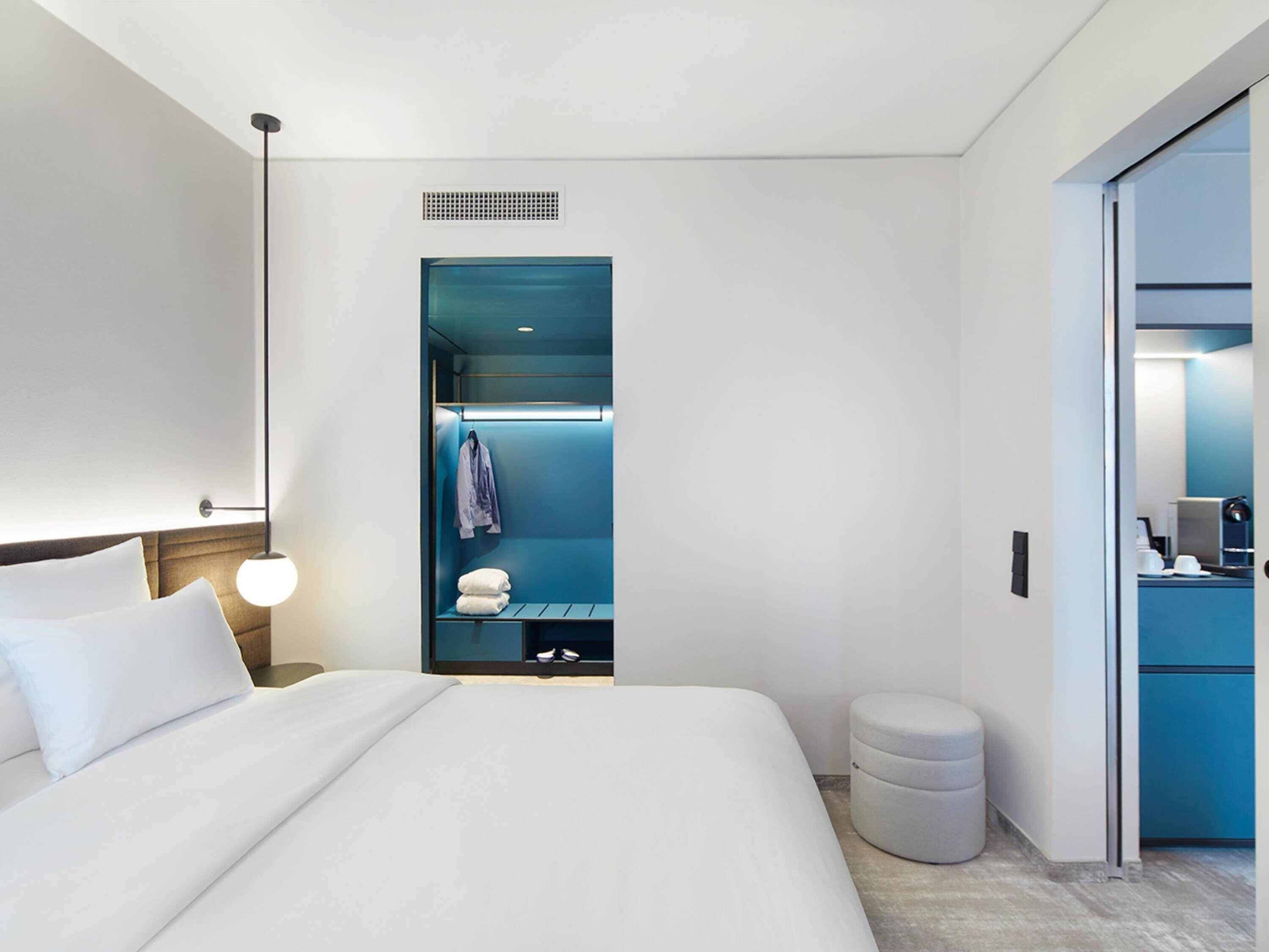 Una habitación de hotel moderna con una cama grande, un vestidor amplio, un reposapiés redondo y un escritorio con una computadora.