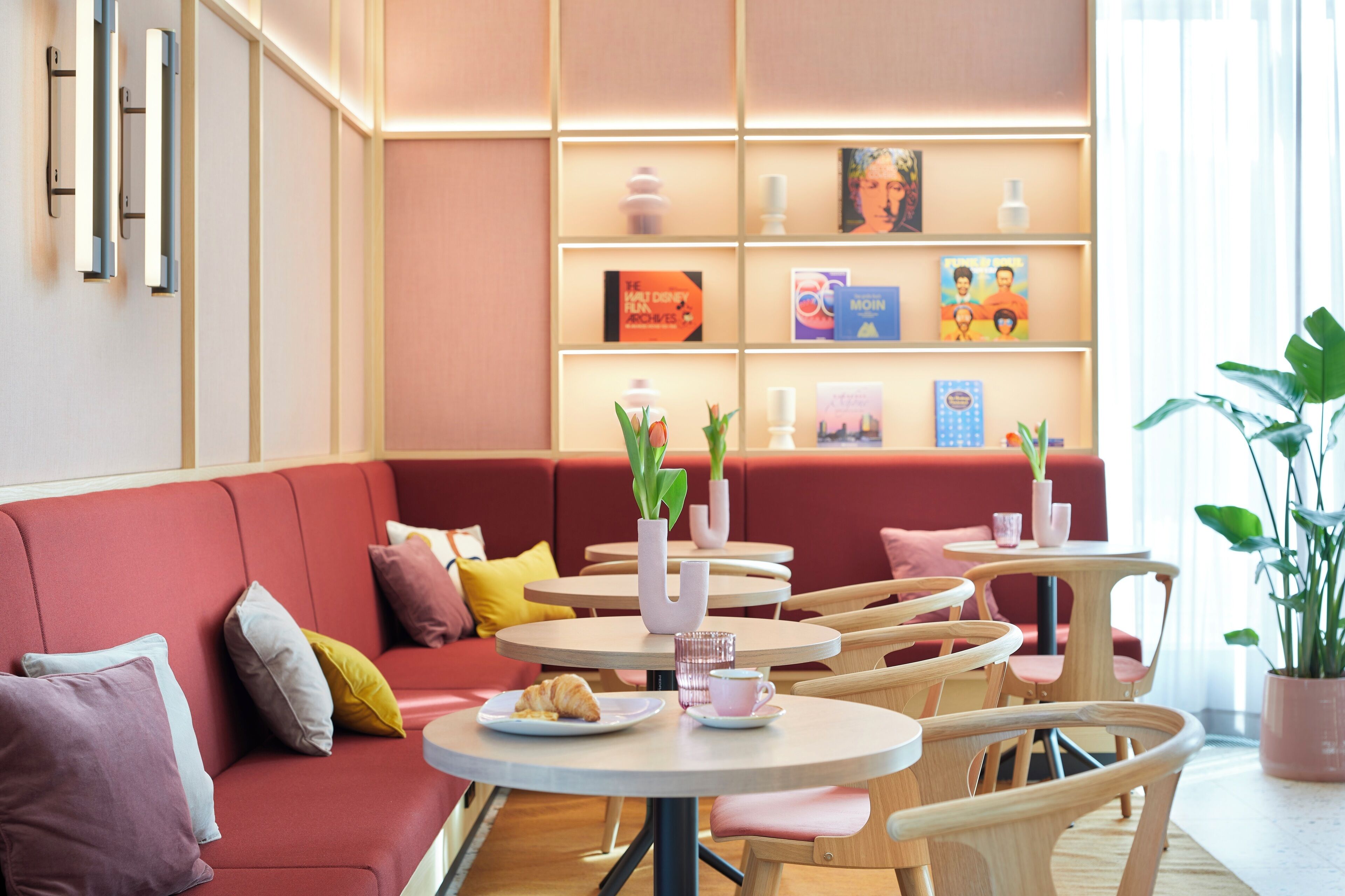 Een modern café met rode zitplaatsen, ronde tafels en diverse boeken en decoratieve objecten op planken.