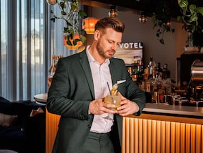 Ein Mann im dunklen Anzug hält einen Cocktail mit Trinkhalm, im Hintergrund ist ein Barbereich mit einem Schild 'VOTEL' zu sehen.